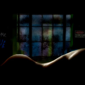 Dark, atmospheric self-portrait of a nude figure reclining in shadow, with a narrow band of light tracing the curves of the body. Behind the figure, textured blue-green windowpanes glow faintly. On the wall, the words “Become” and “Love” appear opposite a sign reading “Warning: Do Not Touch,” creating a tension between intimacy, vulnerability, and distance.