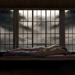 Wide-format image of a nude figure lying face down on a cushioned window seat inside a dark wooden cabin. The figure is seen from behind, stretched horizontally across the lower portion of a large wall of multipaned windows. Beyond the glass is a misty midwinter North Cascades landscape, with snow-covered slopes, dark evergreen forests, and distant mountains emerging through layers of fog and low cloud. The pale landscape and softly illuminated body contrast with the deep brown and black shadows of the cabin interior.